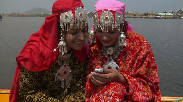 印度查谟和克什米尔的达尔湖上，两名妇女穿着传统服装，一边用手机发短信，一边身后漂浮着墨镜视频素材