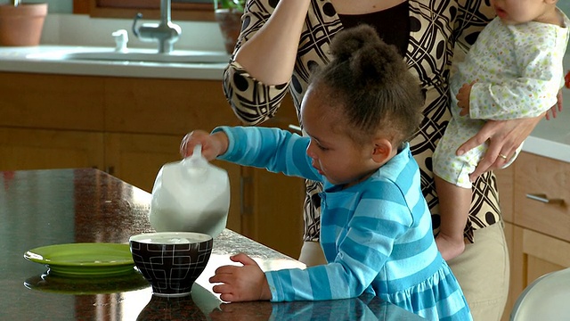 美国弗吉尼亚州里士满的TU女孩把牛奶倒在碗里并洒了出来，而母亲抱着婴儿在讲电话视频下载