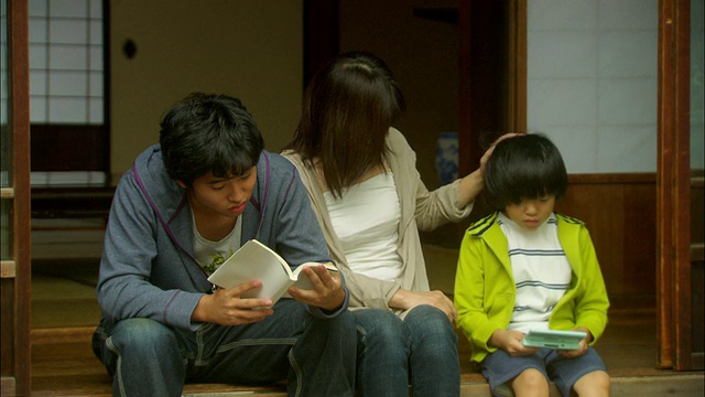 在日本东京，母亲和一个十几岁的男孩在读书，一个小男孩在玩电子游戏，并排坐在门口视频下载