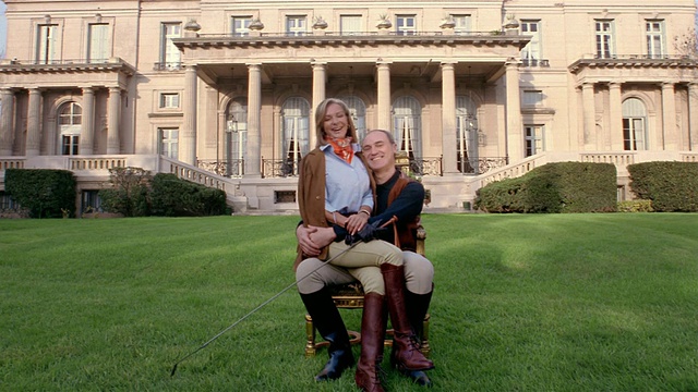 一对富有的骑马夫妇坐在豪宅前的椅子上(女人坐在男人的腿上)的广角照片视频素材