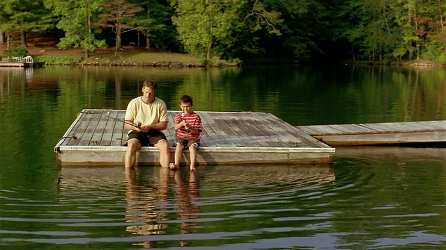 长镜头父子俩扛着鱼竿到码头的尽头/脚浸在水里坐在边上视频素材