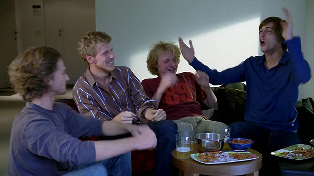 中镜头拍摄了四名坐在沙发上看电视的年轻人，他们在欢呼/鼓掌+唱歌视频素材