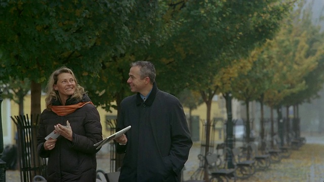 慢镜头中景拍摄男人和女人走过公园长椅+树木与秋叶/布拉格视频下载