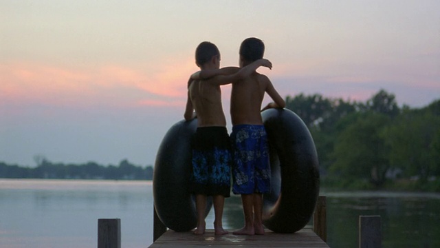 黄昏时分，两个男孩带着内胎在码头上看着池塘，他们互相抱着胳膊视频下载