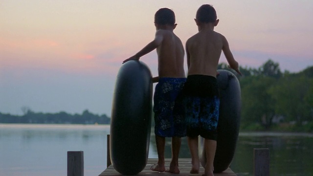 后视图两个男孩滚动内胎下码头在黄昏/长池，纽约视频下载