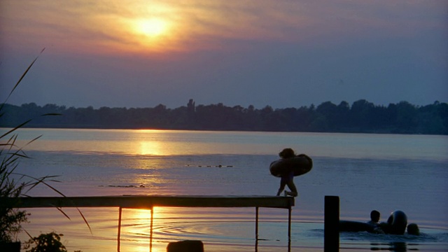 慢镜头:夕阳西下，三个带着内胎的孩子从码头上跳入湖中视频下载
