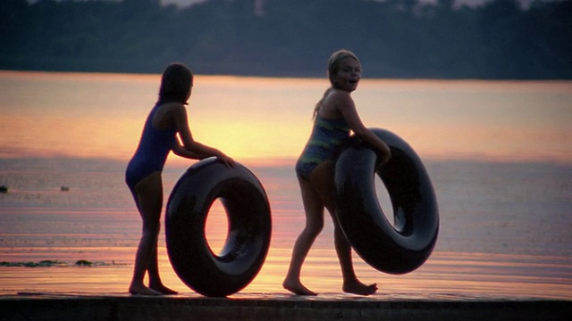 慢镜头:夕阳下，两个女孩在码头上滚动内胎，湖面上倒映着五彩缤纷的天空视频下载