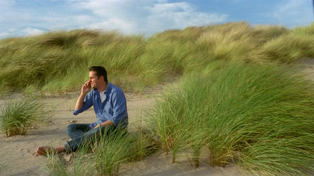 宽镜头拍摄横跨草地沙丘，过去的人坐在沙子上，并在大风天讲手机视频下载