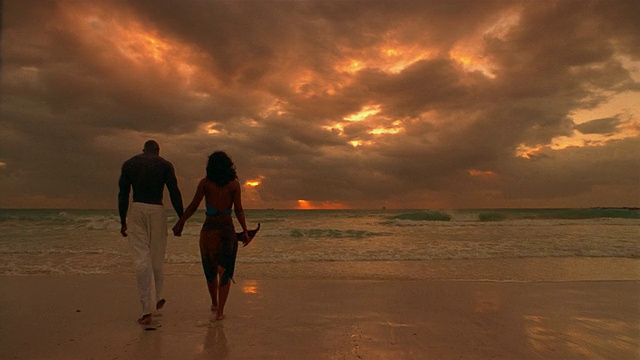 慢镜头宽镜头情侣牵手从水边沿着海滩走向相机/日落/停止和亲吻/走回水里视频下载