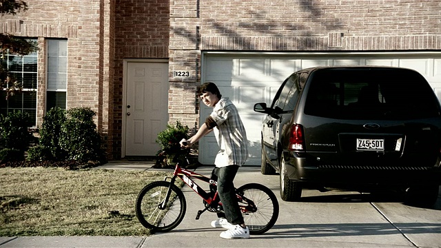 中等镜头的少年骑着自行车/停在郊区的房子前，看着街对面的摄像头视频素材