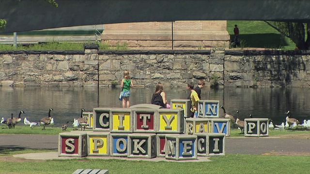 美国华盛顿州斯波坎市，孩子们在河边的大型建筑群雕塑旁喂鹅视频下载
