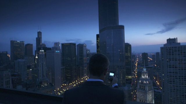 美国伊利诺斯州芝加哥，暮色中，在屋顶上使用手机的商人的后视图，摩天大楼的背景视频素材