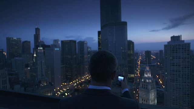 美国伊利诺斯州芝加哥，暮色中，在屋顶上使用手机的商人的后视图，摩天大楼的背景视频素材