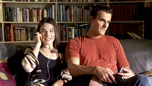 美国加州谢尔曼奥克斯，一对年轻夫妇坐在客厅沙发上看电视，妻子在用手机。视频素材
