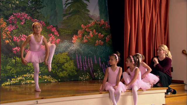 中等镜头的年轻芭蕾舞演员在舞台上跳舞/其他女孩和老师坐在舞台边缘观看/鼓掌的芭蕾舞演员行屈膝礼视频素材