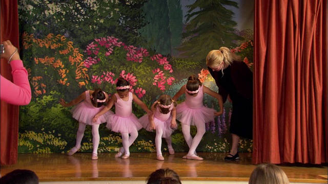 广角镜头四个穿着芭蕾舞短裙的年轻女孩在舞台上微笑，老师/女孩们在行屈膝礼，失去平衡/女孩们在观众面前拍照视频下载