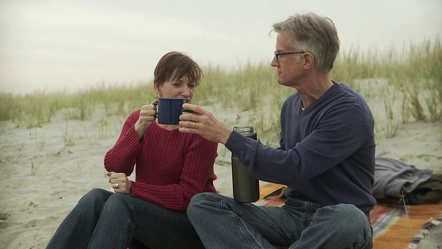在美国新泽西州的海滩上，一对成年夫妇坐在毯子上倒咖啡喝咖啡视频下载