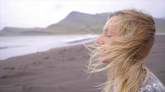 年轻女子站在黑色的沙滩上凝望大海，头发随风飘动——冰岛——慢镜头——人们旅行的自然概念视频素材