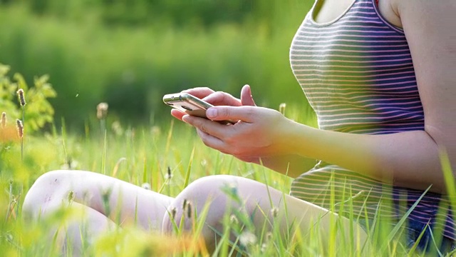 快乐的年轻女子坐在绿色草坪上，在日落背景下使用智能手机视频素材