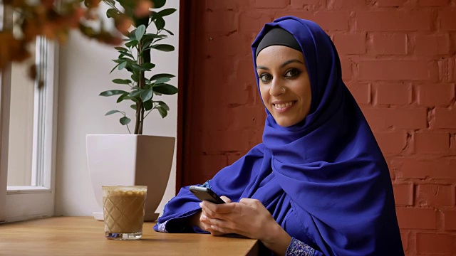 年轻的穆斯林妇女戴着头巾，鼻子穿孔，用手机和微笑的镜头，坐在现代的咖啡馆视频下载