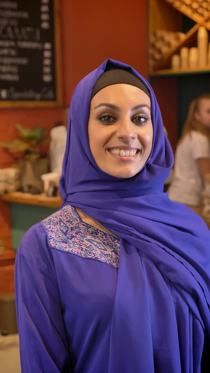 垂直视频拍摄，年轻的穆斯林妇女在镜头前微笑，坐在咖啡馆视频下载