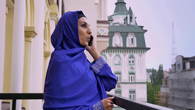 年轻美丽的穆斯林妇女戴着头巾站在阳台上，在雨中打电话视频下载