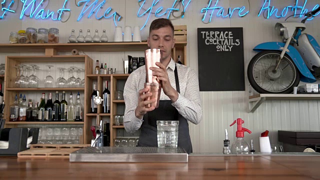 一位英俊的年轻调酒师站在吧台前，用调酒器摇着一杯蓝色的鸡尾酒。然后他把摇酒器里的东西倒进杯子里。视频素材