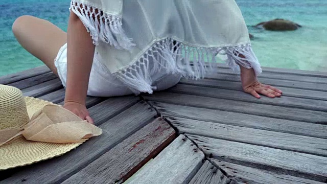 女人戴着海帽她开心地坐着，在木桥上喝着椰子汁，看海边沙滩，云和蓝天是无边的背景，复制空间。视频素材