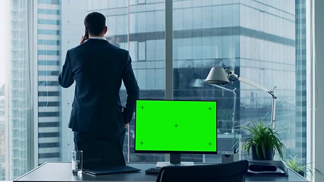 自信的商人在他的办公室打电话，个人电脑在他的桌子上显示绿色的色度键屏幕。在窗口全景大城市景观。视频素材