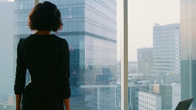 下面的照片是一位成功的女商人穿着醒目的黑色连衣裙走过她的办公室，若有所思地看着窗外。拥有个人电脑和大城市景观的现代商务办公室。视频素材