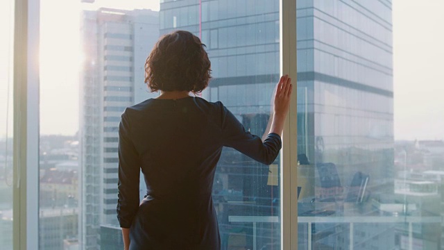 后景拍摄的成功女商人在醒目的黑色连衣裙站在她的办公室，看着窗外，若有所思。拥有个人电脑和大城市景观的现代商务办公室。视频下载