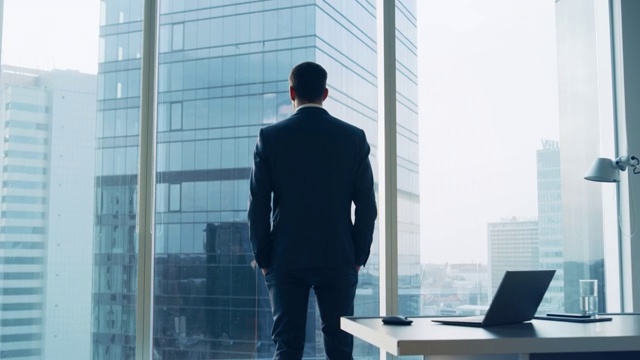 Back View of the Thoughtful Businessman wearing a Suit Standing in His Office, Hands in Pockets and considering Next Big Business Deal, Looking out of the Window.(后视图)大城市商业区全景窗景。视频素材