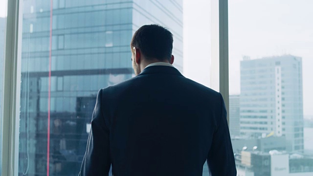 中景拍摄的成功商人穿着西装站在他的办公室，考虑下一个大生意，看着窗外。大城市商业区全景窗景。视频素材