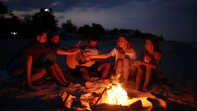 日落时分的沙滩派对，篝火晚会。朋友们围坐在篝火旁，喝着啤酒，唱着吉他。年轻的男男女女拿着装着饮料的玻璃瓶，一边唱一边欢呼视频素材