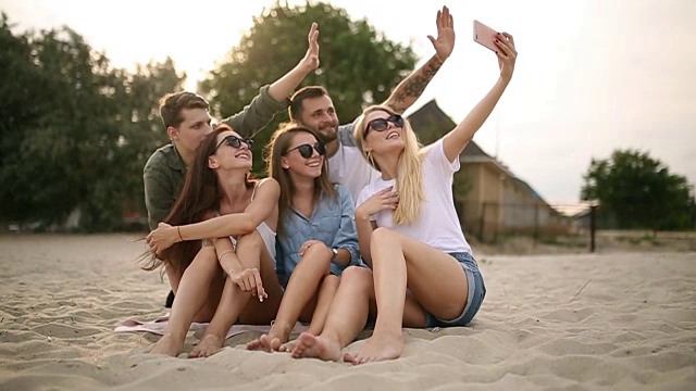 一群年轻的朋友在海滩上自拍。在温暖的夏夜，男男女女坐在沙滩上拍照视频素材