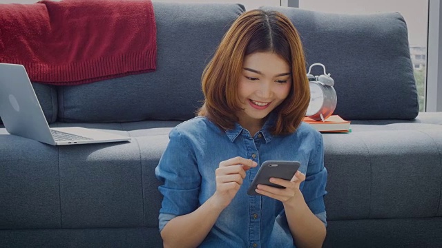 年轻的亚洲女子坐在家里的地板上用手机微笑视频下载