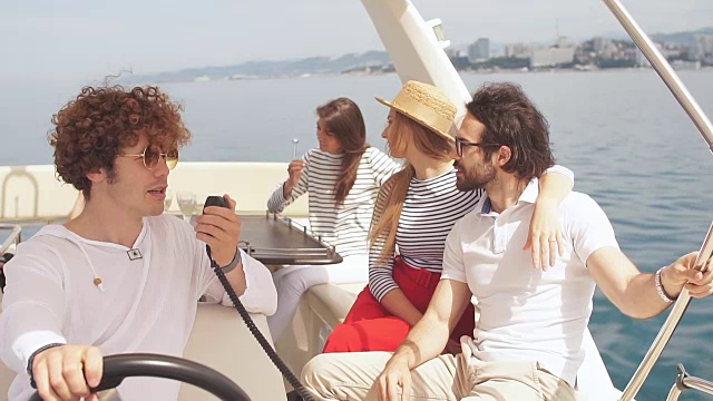 年轻的卷发男人驾驶着一艘海上船，在他快乐的朋友的陪伴下航行，男孩和女孩坐在旁边，享受新鲜的海风视频下载