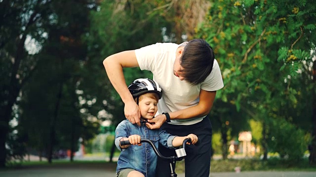 慈爱的父亲给他的小儿子戴上安全帽，然后教他骑自行车，而慈爱的母亲看着他们微笑。视频素材