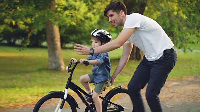 英俊的年轻男子慈爱的父亲正在公园里教他的小儿子骑自行车，在夏天的一天，男孩骑着自行车，爸爸抱着他跑步。视频素材