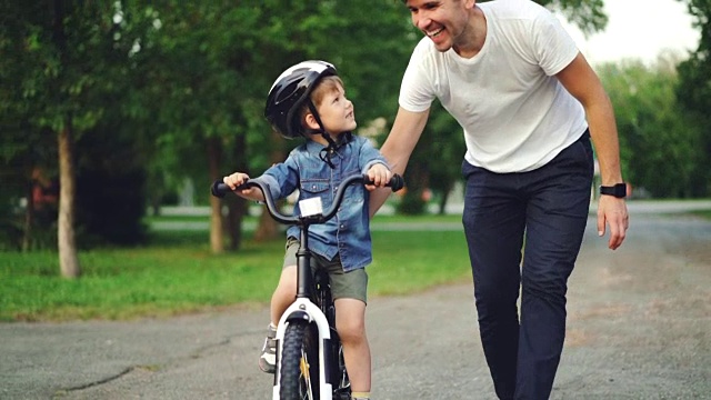 慢镜头，慈爱的父亲教他可爱的儿子骑自行车在公园里，拿着自行车和孩子说话。父亲，童年和积极的生活方式的概念。视频素材