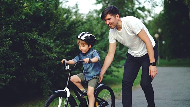 慢动作兴奋的男孩骑自行车和大笑，而他小心的父亲正在帮助他拿着自行车和教孩子骑。家庭、运动和童年的概念。视频素材