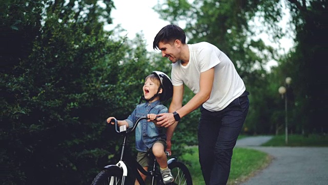 慢镜头:一个快乐的年轻人慈爱的父亲在夏天教他的孩子在绿色的公园里骑自行车，小男孩笑着，喊着，和爸爸一起享受周末。视频下载