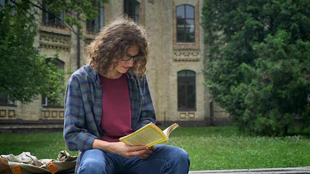 戴眼镜、卷发的帅哥坐在大学附近公园的长椅上看书，认真而专注视频素材