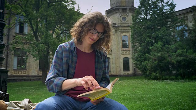 戴着眼镜，一头卷发的年轻帅气的学生坐在大学大楼附近的公园里看书，看着相机视频素材