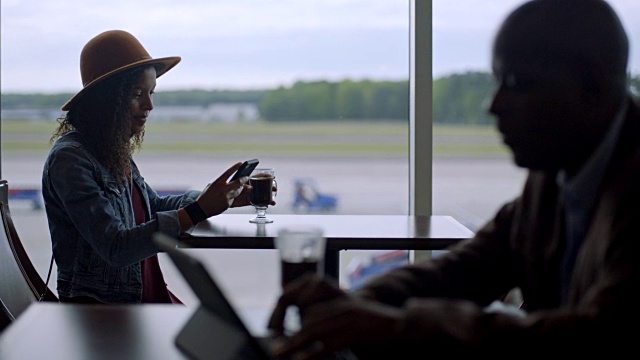 混血的年轻女性旅客一边喝着咖啡，一边在机场候机厅的咖啡馆窗口边等待。视频素材