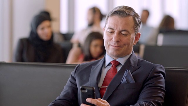 带着智能手机微笑的商人坐在机场门口的等候区。视频素材