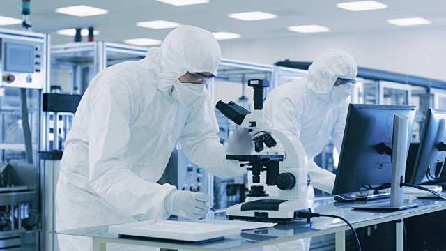 在实验室，科学家穿着防护服做研究，使用显微镜和输入数据到个人电脑。现代制造半导体和制药项目。视频购买