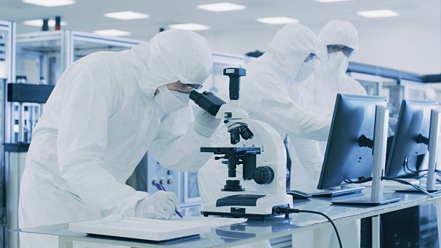 在实验室，科学家穿着防护服做研究，使用显微镜和输入数据到个人电脑。现代制造半导体和制药项目。视频素材