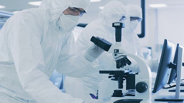 在实验室，科学家穿着防护服做研究，使用显微镜和输入数据到个人电脑。现代制造半导体和制药项目。视频素材