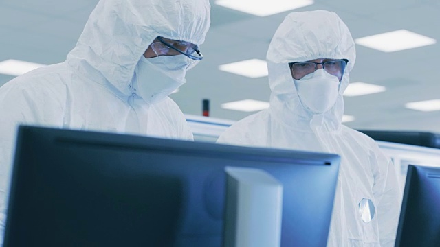 两名穿着无菌服的科学家/技术人员在个人电脑上检查数据，进行讨论。他们在制药制造实验室工作。视频素材
